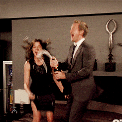 Two characters from How I Met Your Mother jump up and down on a bed in celebration as one sprays a champagne bottle.